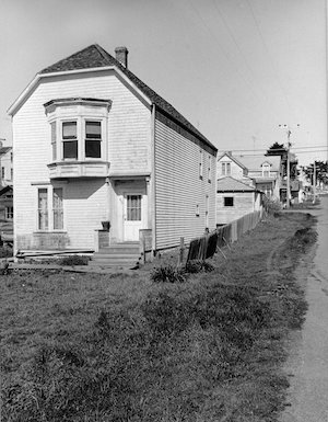 A narrow, two-story building in a grassy lot with buildings behind it