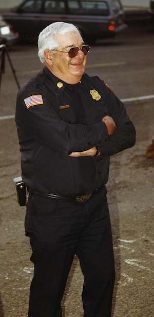 A smiling man with white hair wearing sunglasses and fire department uniform standing with arms crossed