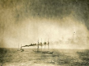 A steam boat towing a larger boat with four masts in the water