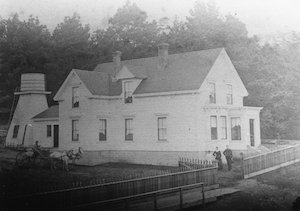 View of a two-story home with water tower in the back. A family of three stand in the yard behind a fence.