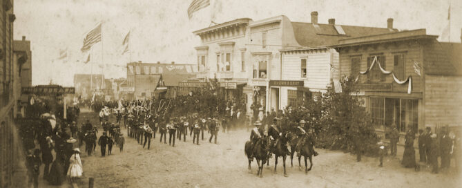 Uniformed men mounted on horses advance eastward on Main Street, followed by a marching band and then ranks of other walking people. Above the street are many large flags waving in the breeze atop very tall "liberty poles.” Both sides of the street are lined with people standing on boardwalks.