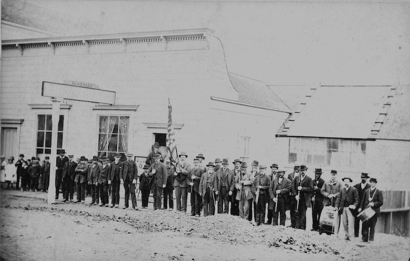 A line of men pose in front of a building holding rifles at salute position