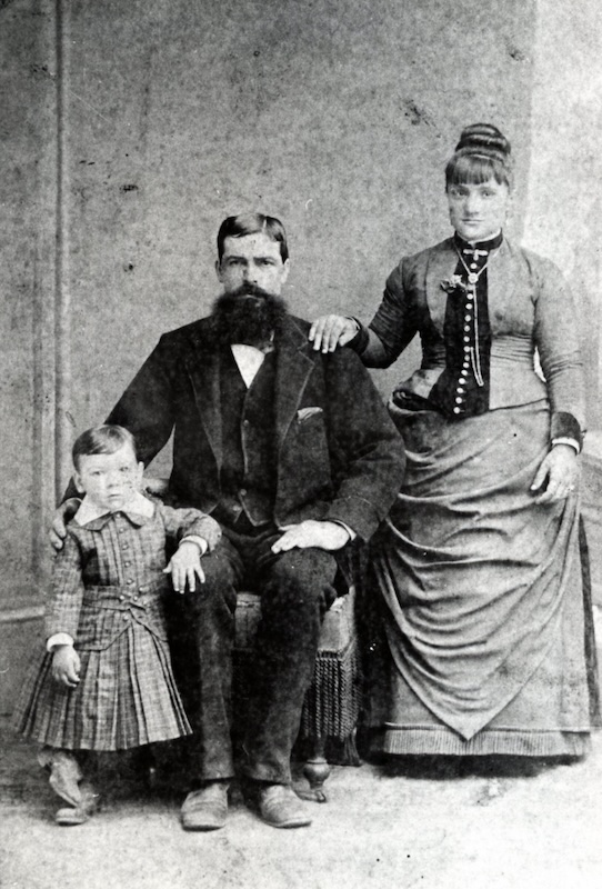Small child standing next to seated man. A woman stands behind the man.
