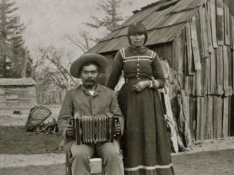 Man seated with woman standing beside him in front of cabin