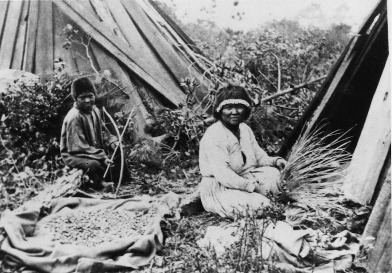 A young boy with a bow and arrow sits behind a Native American basket maker.